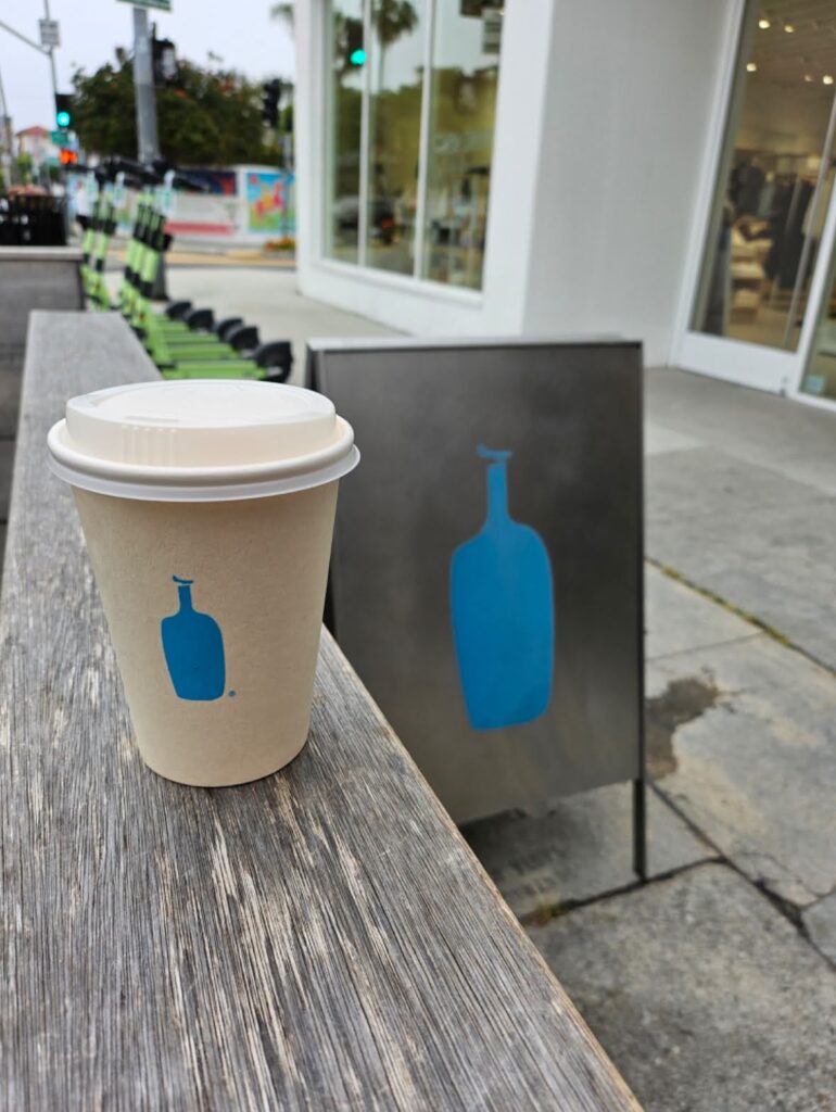 A cup of blue bottle coffee in front of the blue bottle coffee sign in Venice Beach