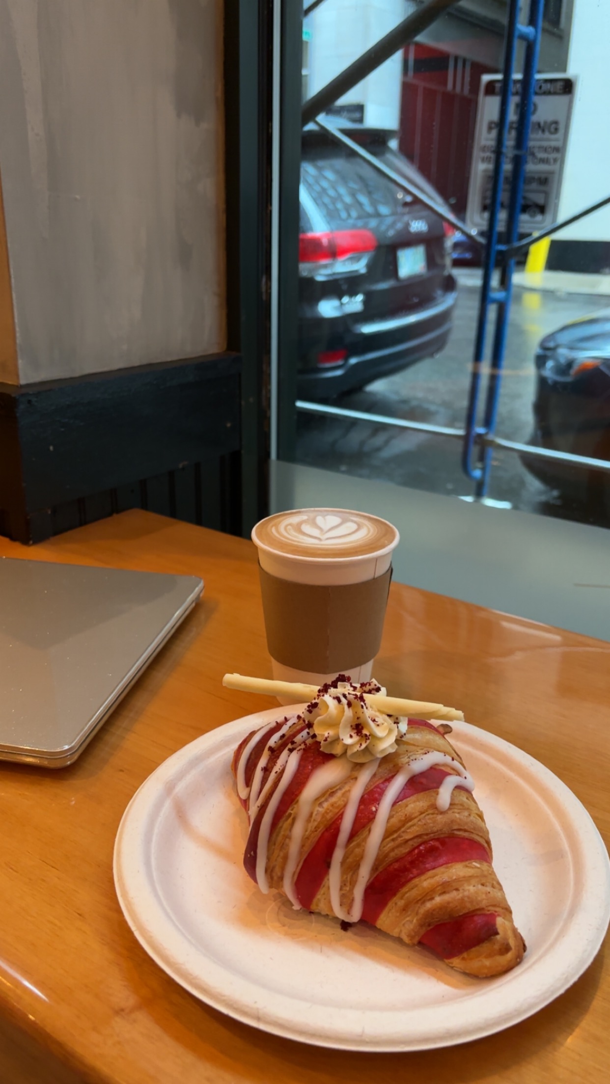 A red velvet croissant and a coffee with a computer sitting nearby overlooking a window