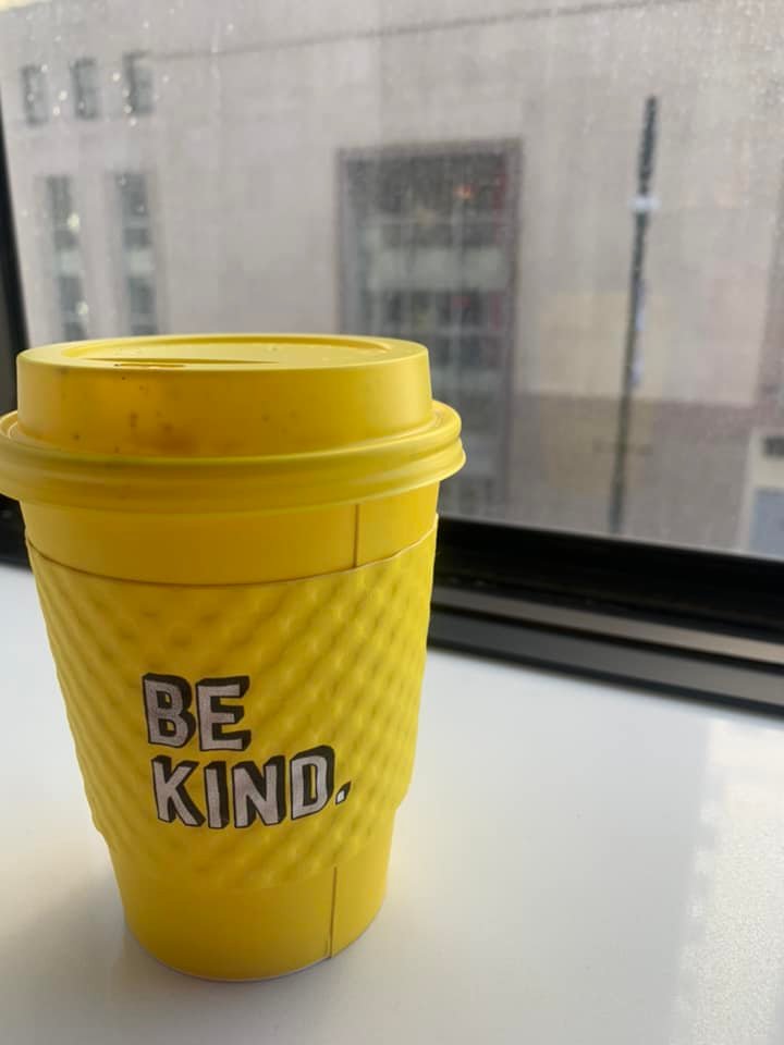 A yellow hot coffee cup with a sleeve that says "be kind"