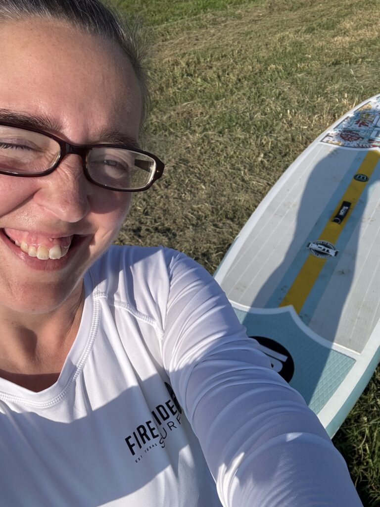 A woman taking a selfie with a stand up paddle board in the background.