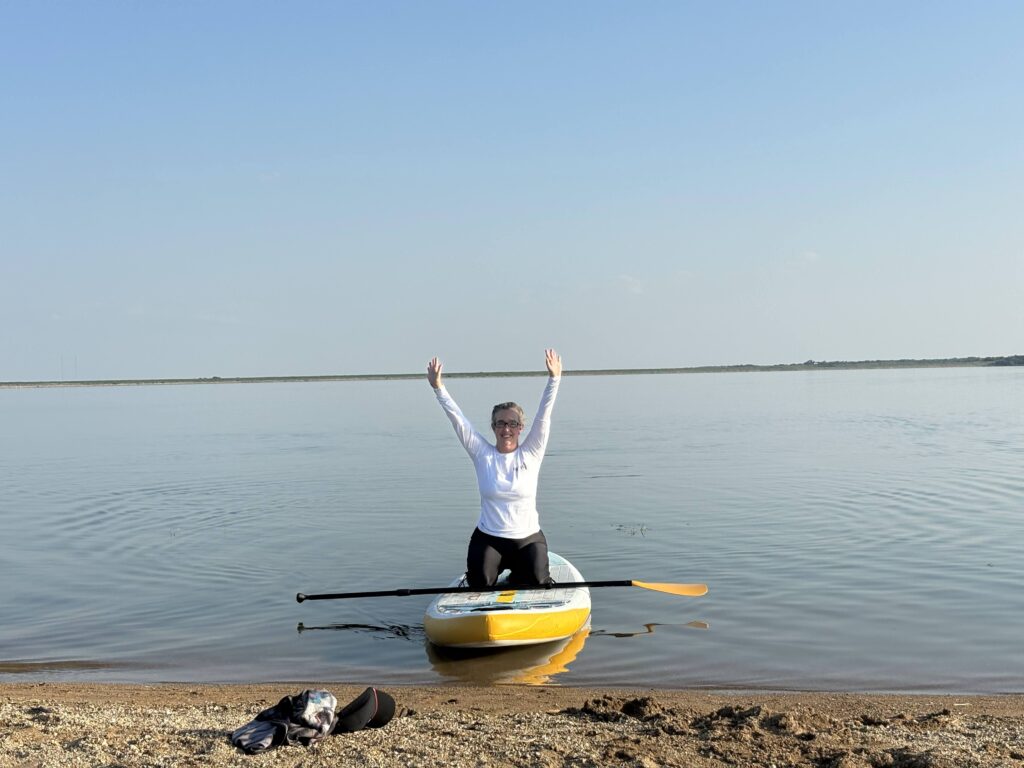 A woman on her knees in a stand-up paddle board on the water with her hands in the air