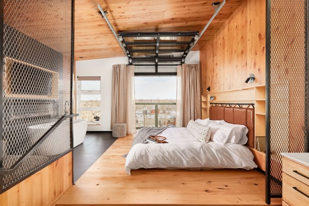 A hotel room at Kingship Landing with wood around it, a bed, a garage door with windows up and a bathtub on the other side of the bed