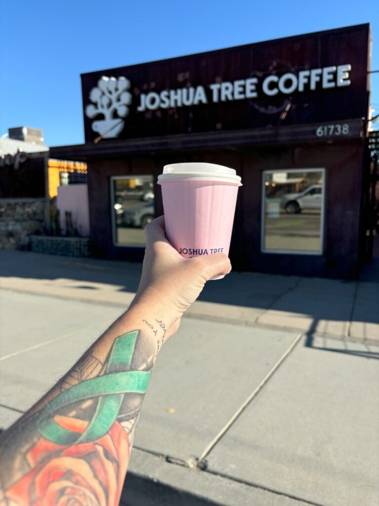 An arm with a tattoo holding a pink coffee cup with a black building that says "Joshua Tree Coffee" in the background